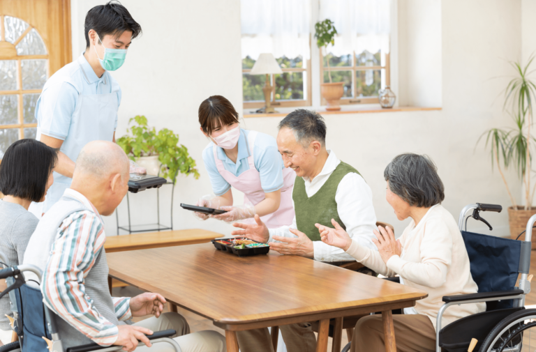 介護施設の食事時間。スタッフが入居者に食事を提供している共有食堂風景。